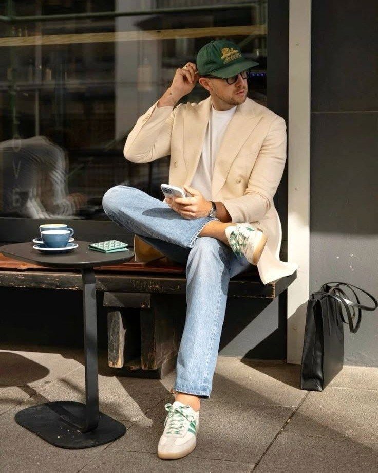 Man wearing a beige blazer, light blue jeans, and white sneakers sitting outdoors.