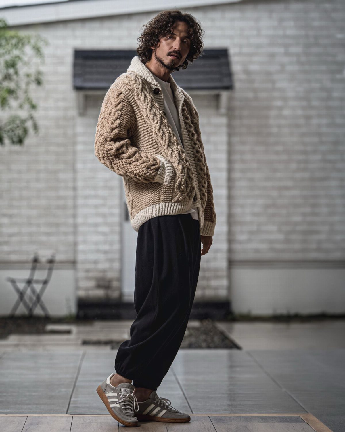 Man wearing a beige cable-knit cardigan, black trousers, and grey Adidas Samba sneakers.