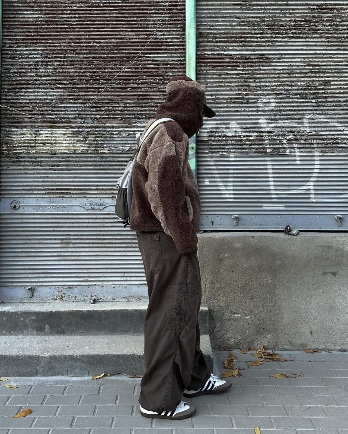 Person wearing a brown fuzzy outfit and black Adidas Samba sneakers by a shutter.