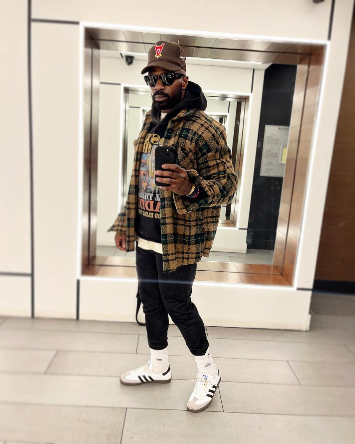 Man in plaid jacket, black hoodie, and white Adidas Samba sneakers taking a selfie.