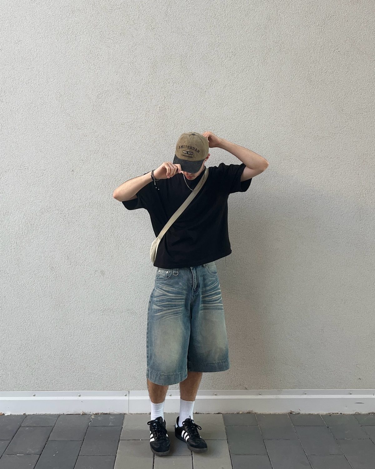 Person wearing black t-shirt, denim shorts, and black Adidas Samba sneakers against grey wall.