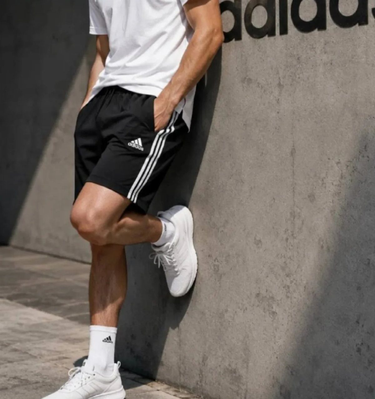 Man in white t-shirt and black Adidas shorts leaning against concrete wall