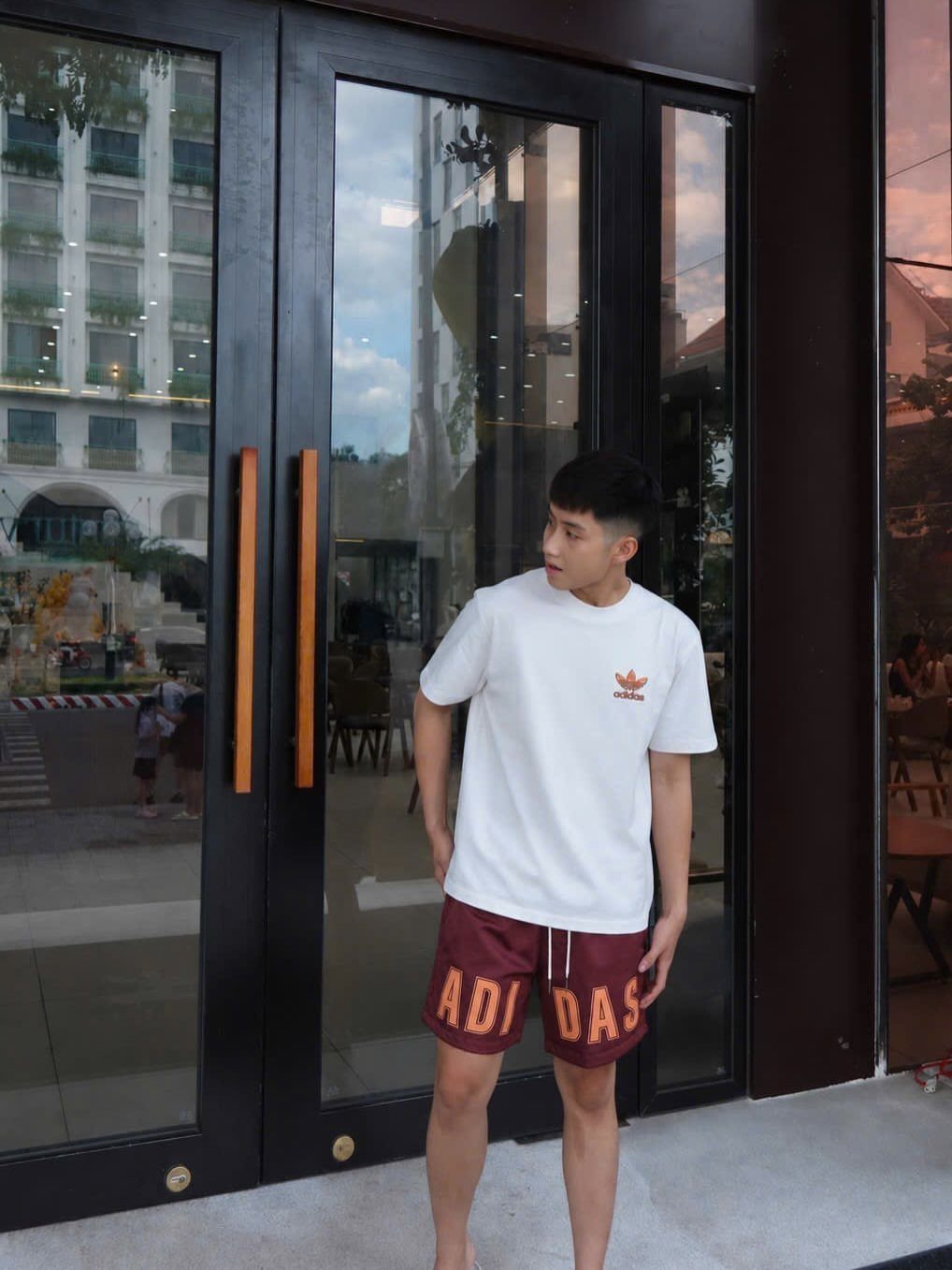 Man wearing a white t-shirt and maroon Adidas shorts standing by glass doors.