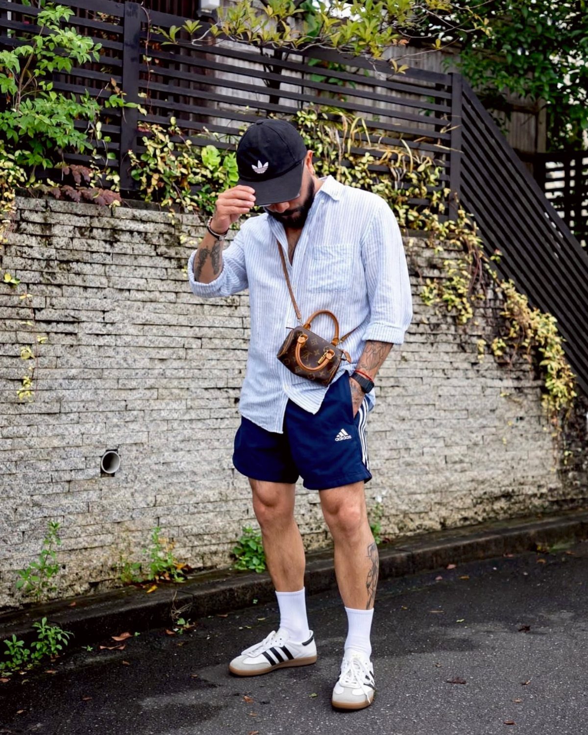 Man in navy Adidas shorts, striped button-down shirt, and white sneakers posing outdoors.