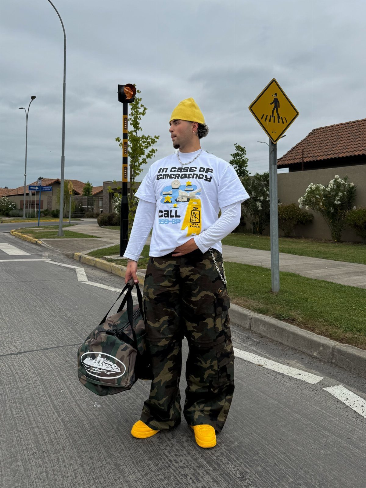 Man in camouflage cargo pants, white graphic tee, and yellow accessories on a street.