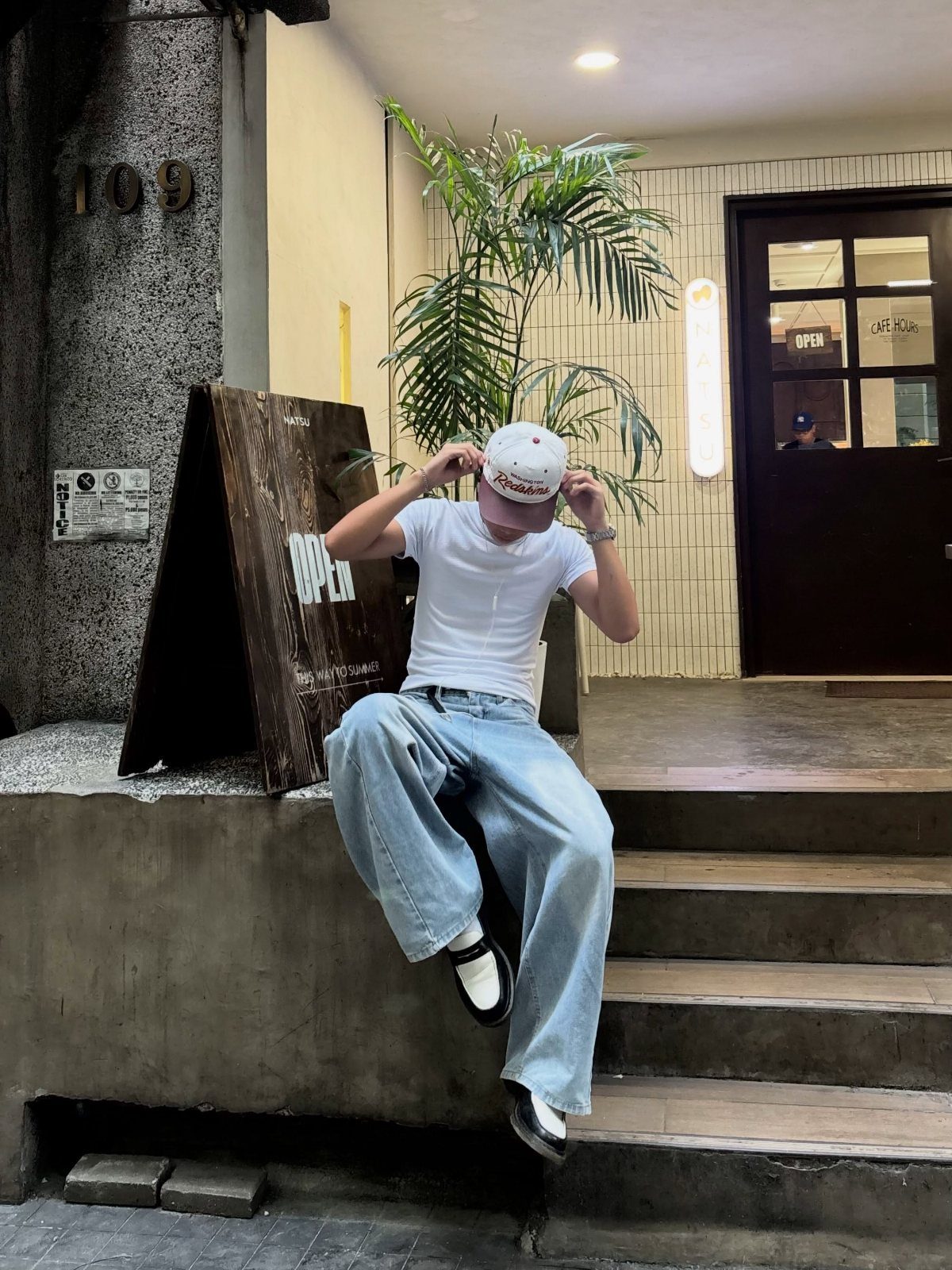 Man in a fitted white t-shirt and baggy light blue jeans sitting outdoors.