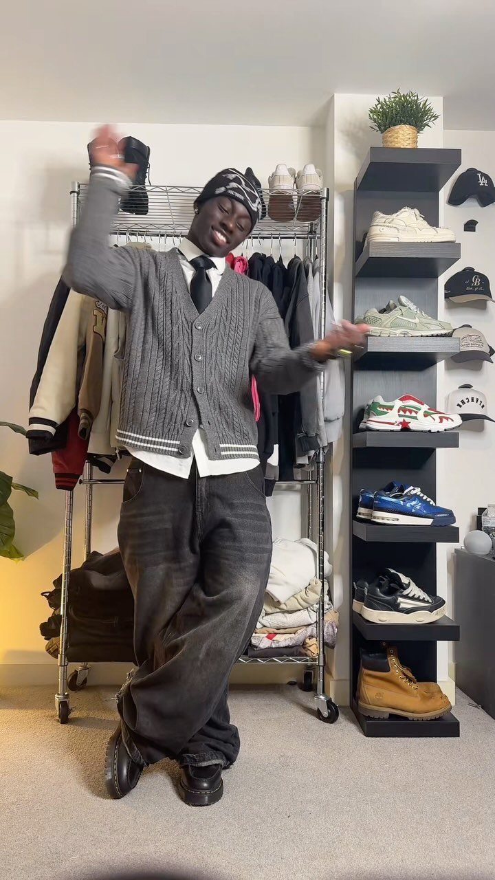 Man in grey cardigan and baggy dark grey jeans posing in a bedroom.