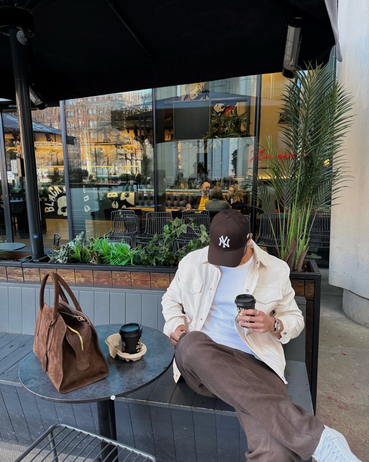 Man wearing a brown baseball cap and baggy outfit sitting at a cafe.