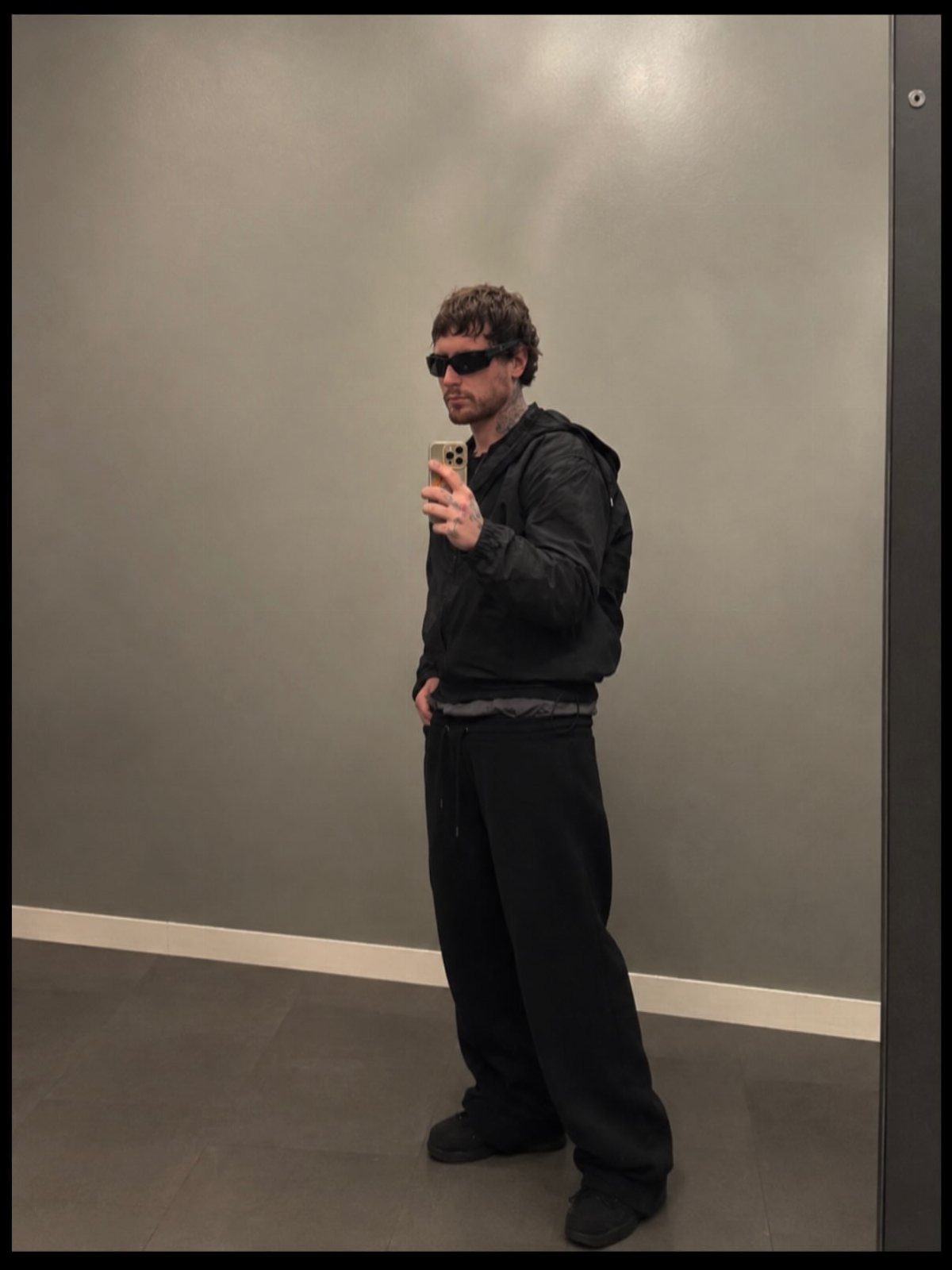 Man in black sunglasses, black windbreaker, and baggy black sweatpants posing for selfie.