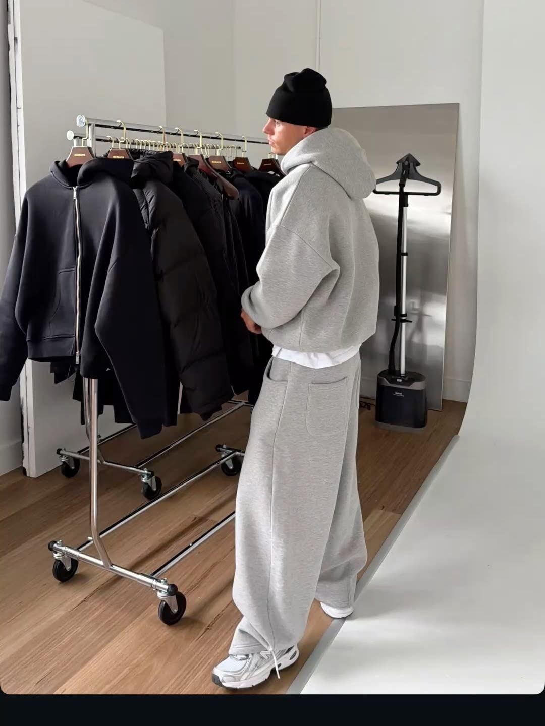 Man in oversized grey hoodie and baggy sweatpants standing by a clothing rack.