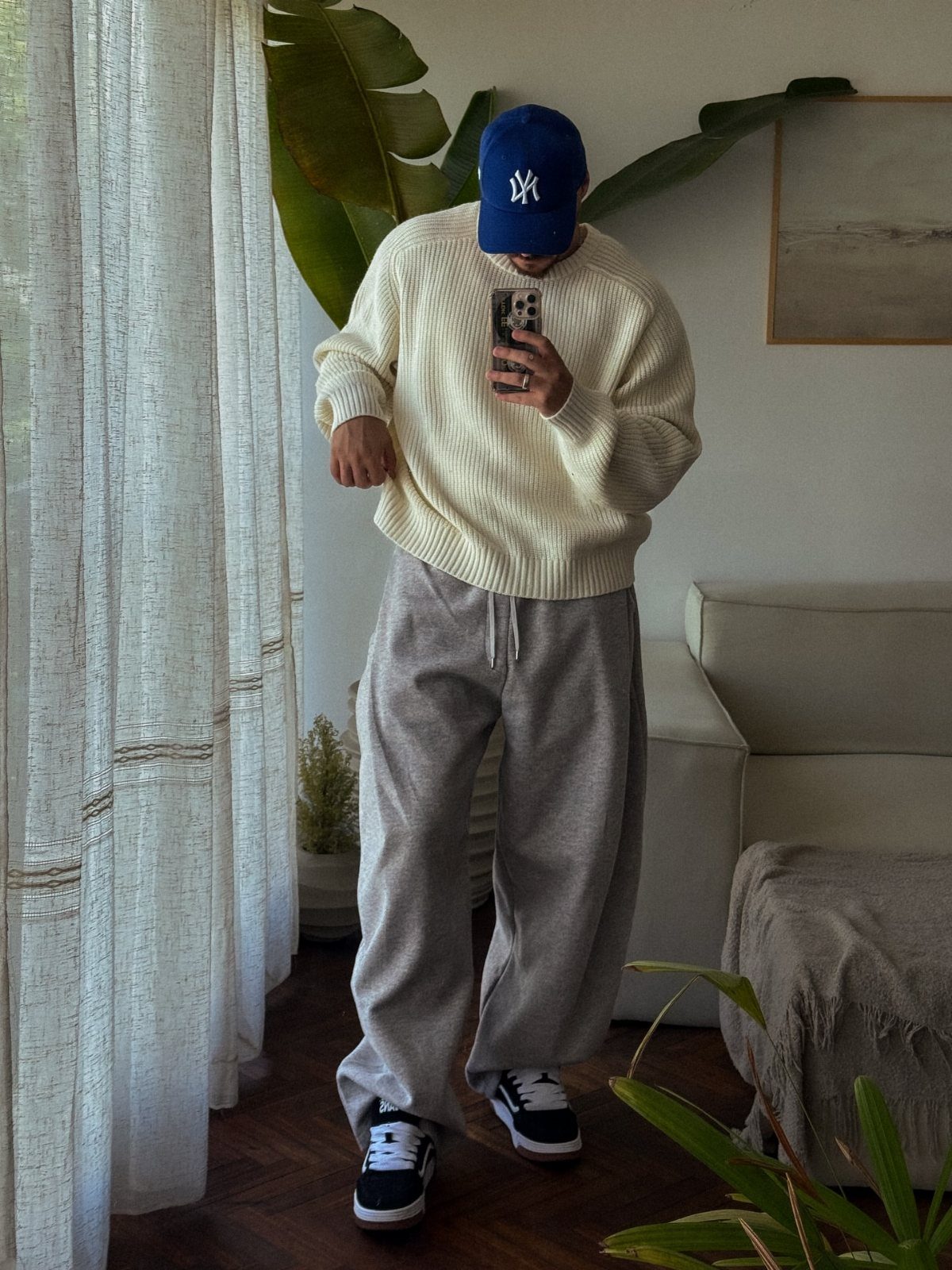 Man wearing a cream knit sweater, baggy grey sweatpants, and blue baseball cap.