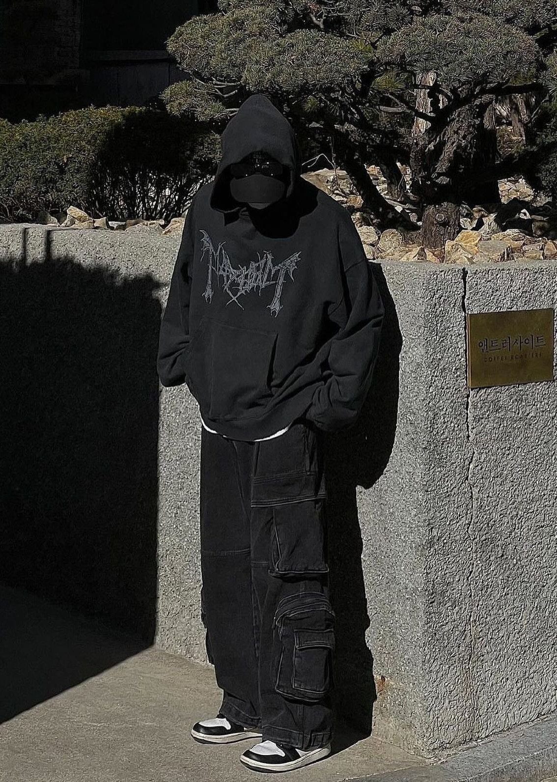Person in oversized black hoodie and baggy black cargo pants standing by stone wall