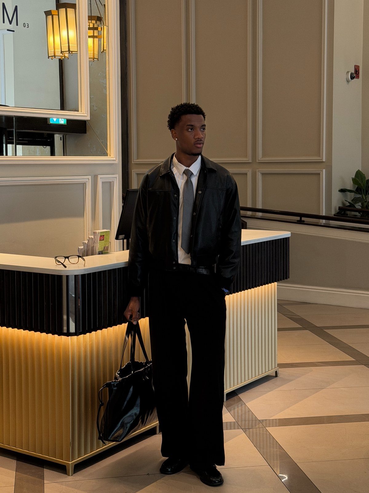 Man in black leather jacket and wide-leg trousers standing in a lobby
