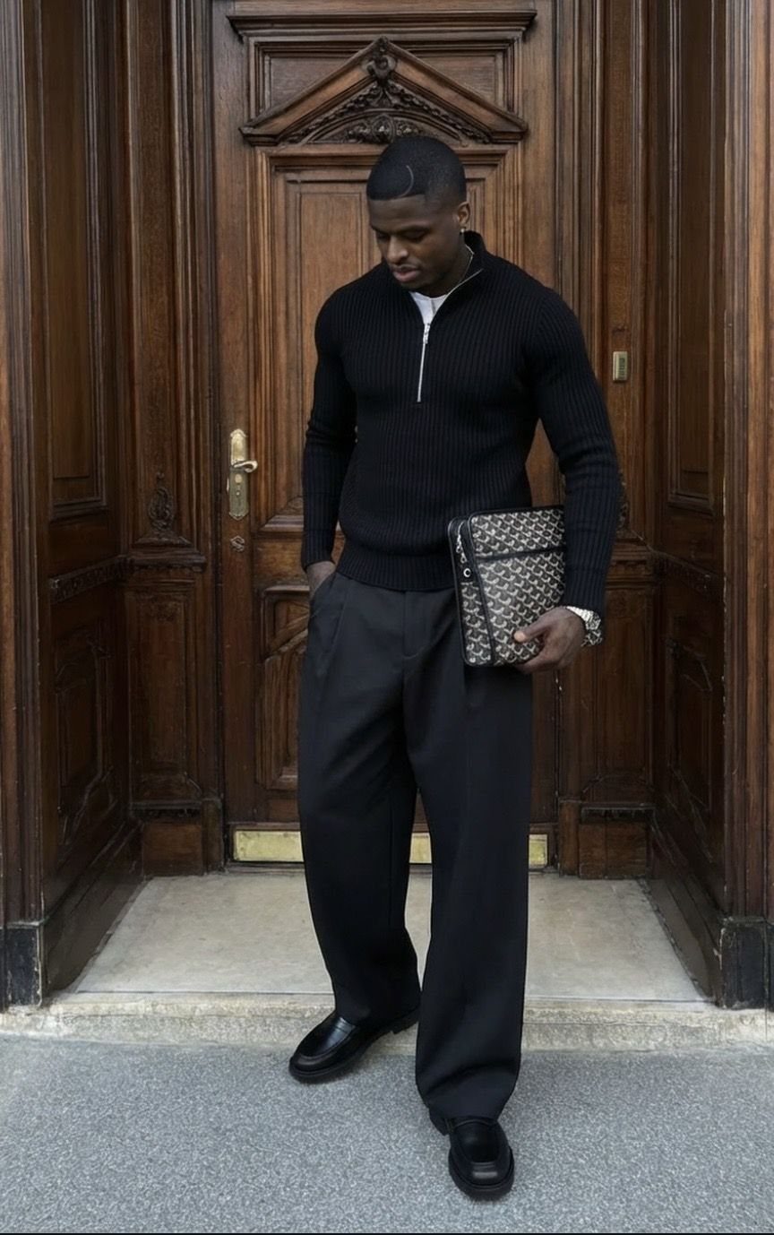 Man in black ribbed sweater and wide-leg trousers standing by wooden door