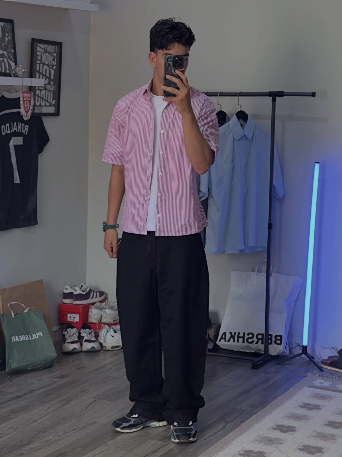 Young man wearing a pink striped shirt and baggy black sweatpants in a room.