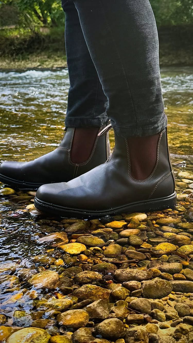Black Chelsea boots and dark jeans standing on river stones in shallow water