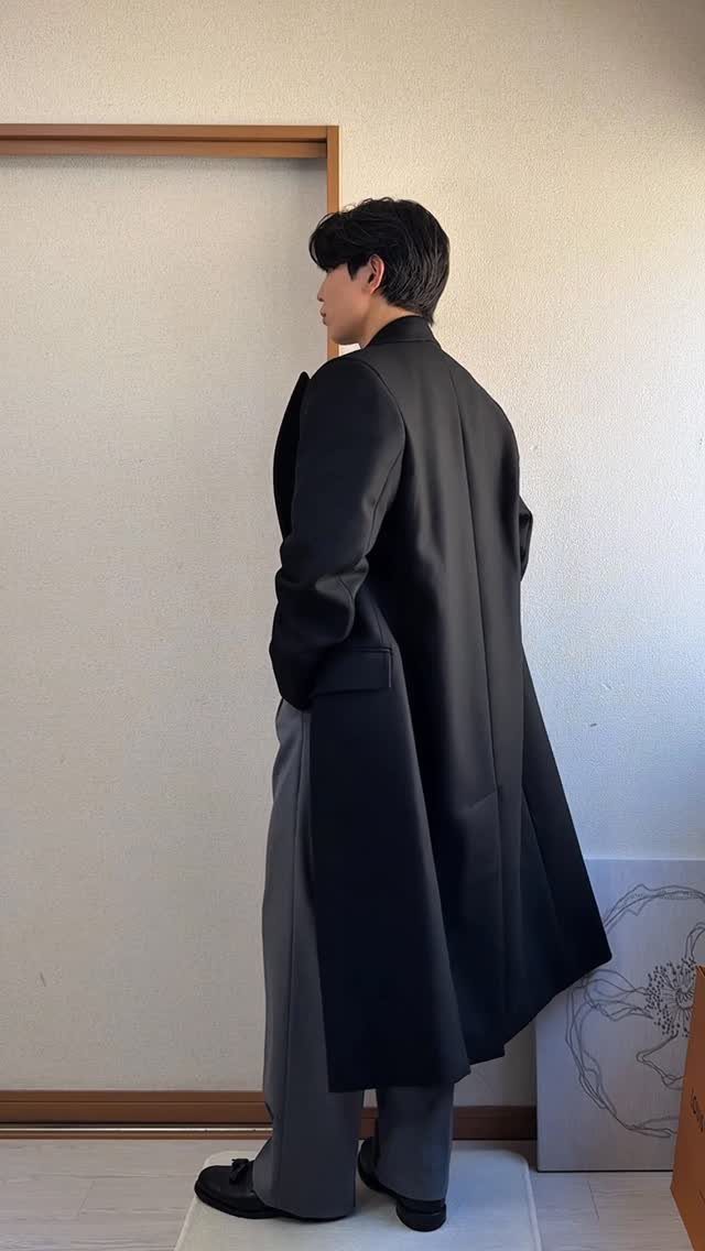 Man wearing a long black overcoat, grey trousers, and black boots standing sideways.