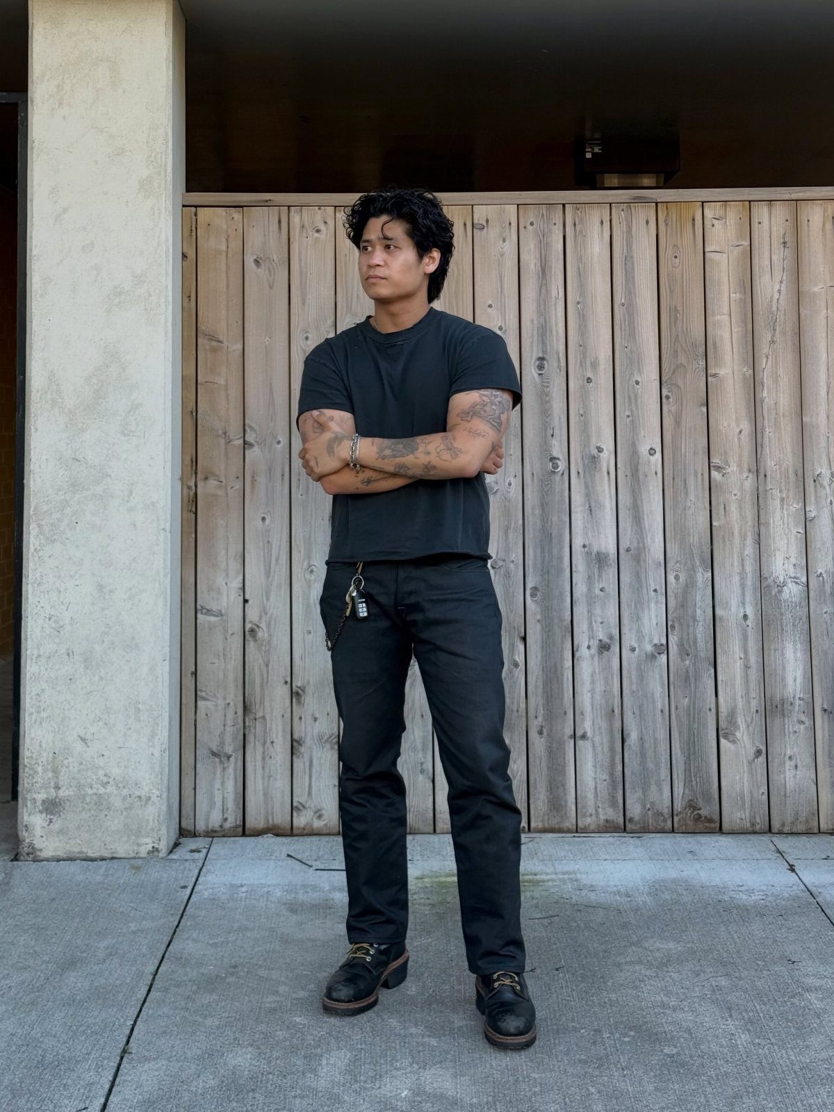 Man wearing a black t-shirt, black pants, and black boots by a fence