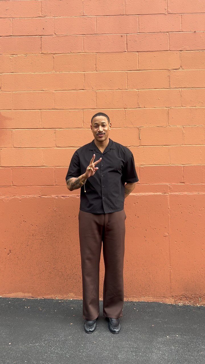 Man in black shirt and brown trousers wearing black loafers against brick wall