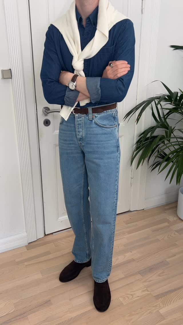 Person wearing light blue jeans, blue shirt, and black loafers standing indoors.