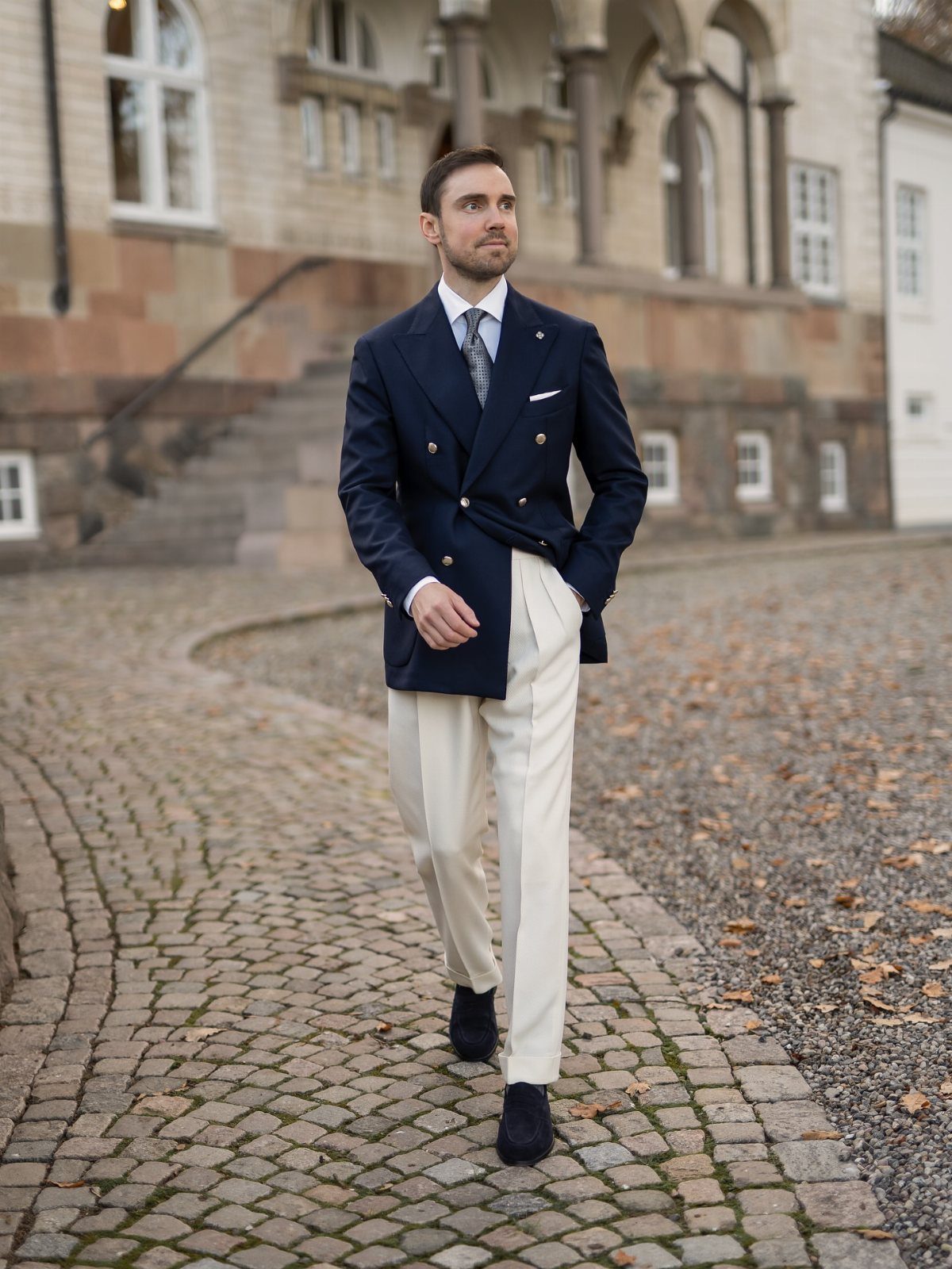 Man in navy blazer and ivory trousers wearing black loafers on cobblestone street