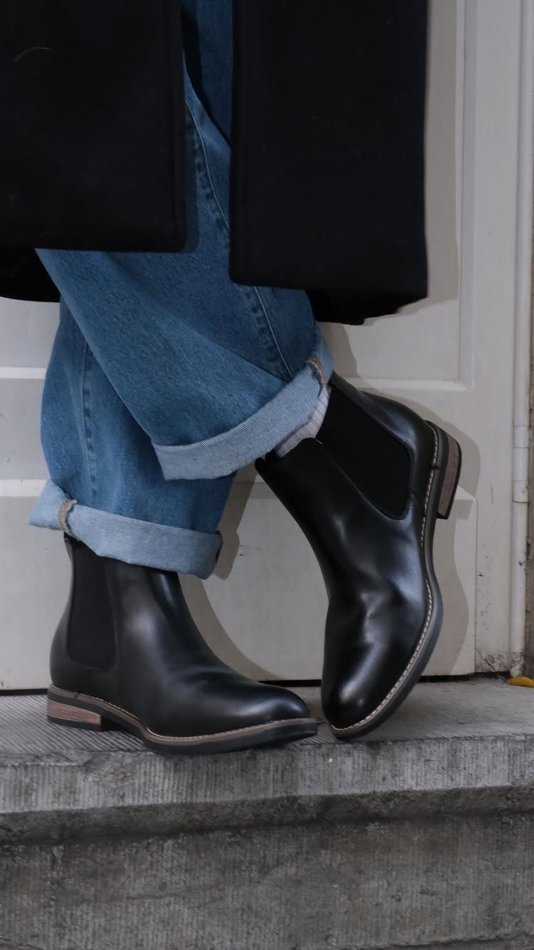 Black leather Chelsea boots paired with cuffed blue jeans and a black coat.