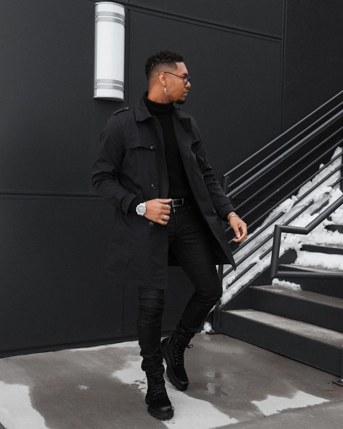 Man in all-black outfit wearing combat boots and skinny jeans by snowy stairs