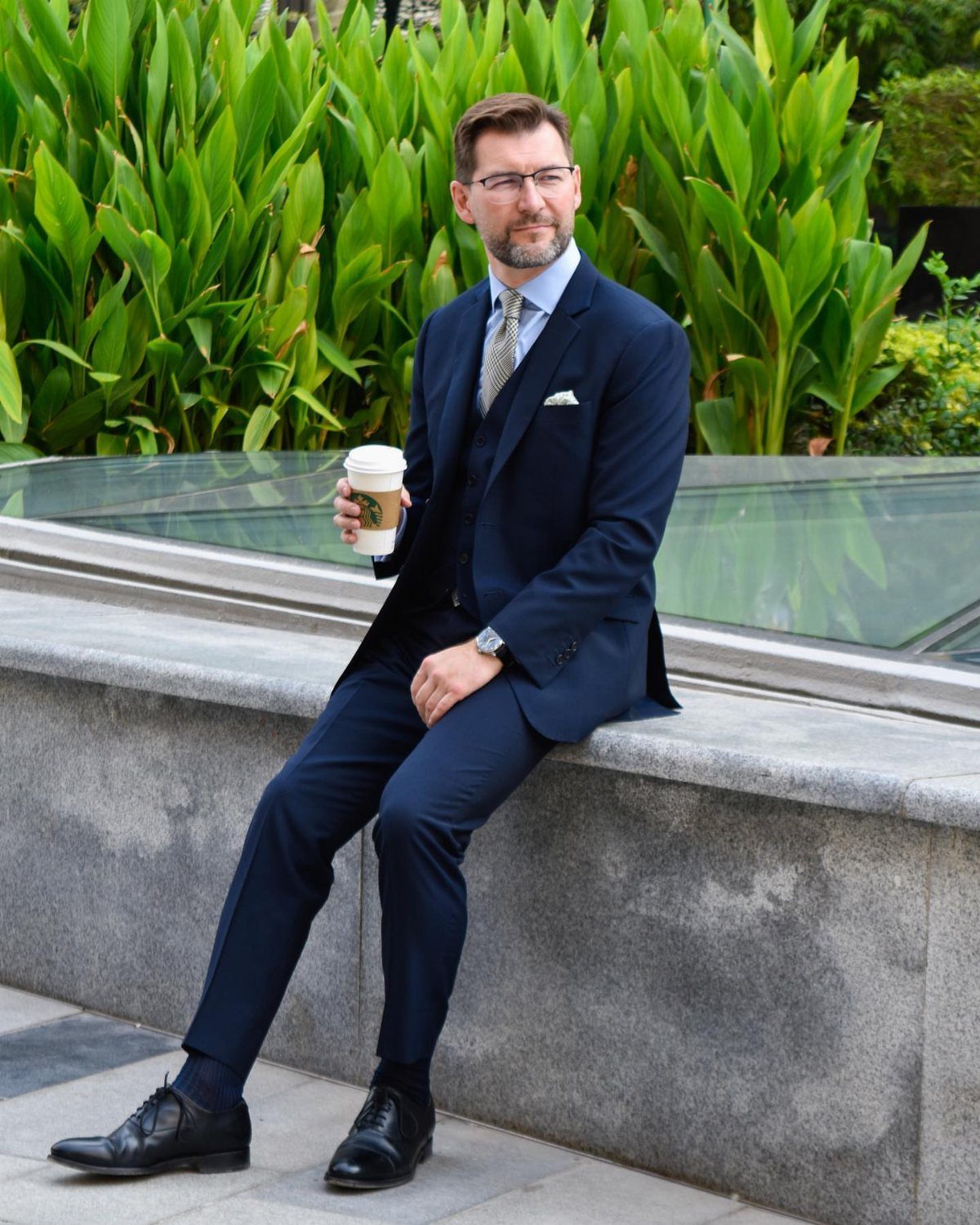 Man in navy blue three-piece suit wearing black leather oxford shoes sitting outdoors.
