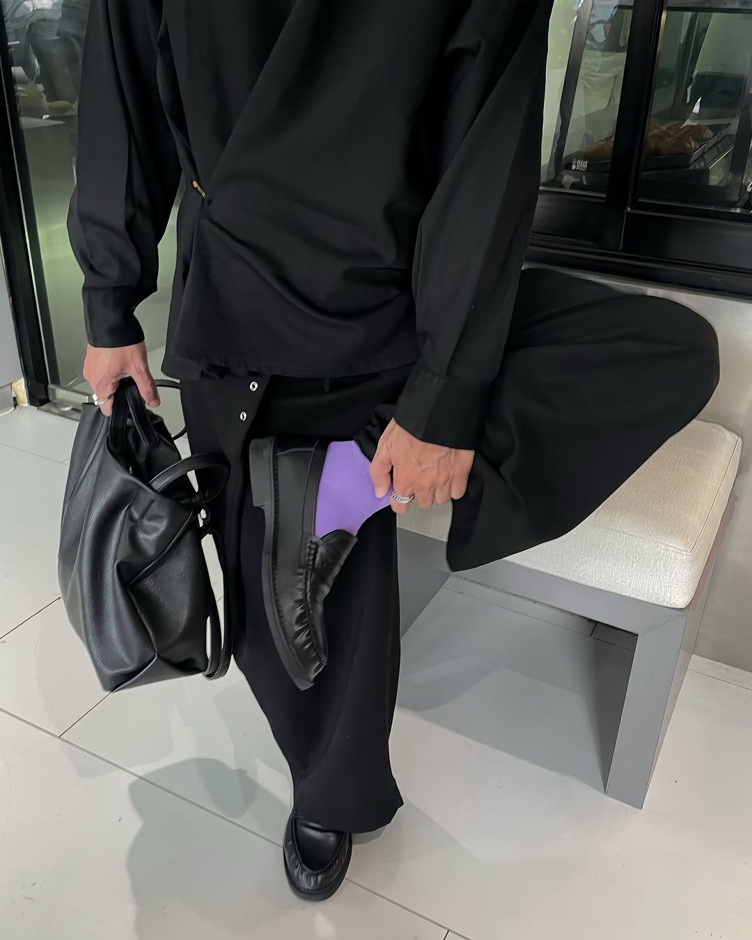 Person in all-black outfit with wide-leg trousers and black leather loafers sitting on bench.