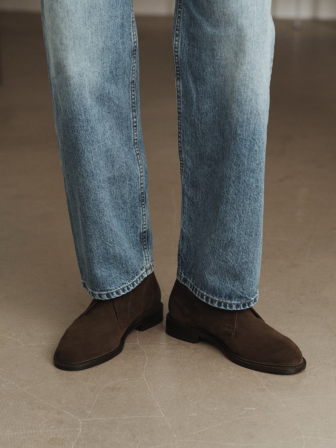 Dark brown suede chukka boots paired with light-wash blue denim jeans on concrete.