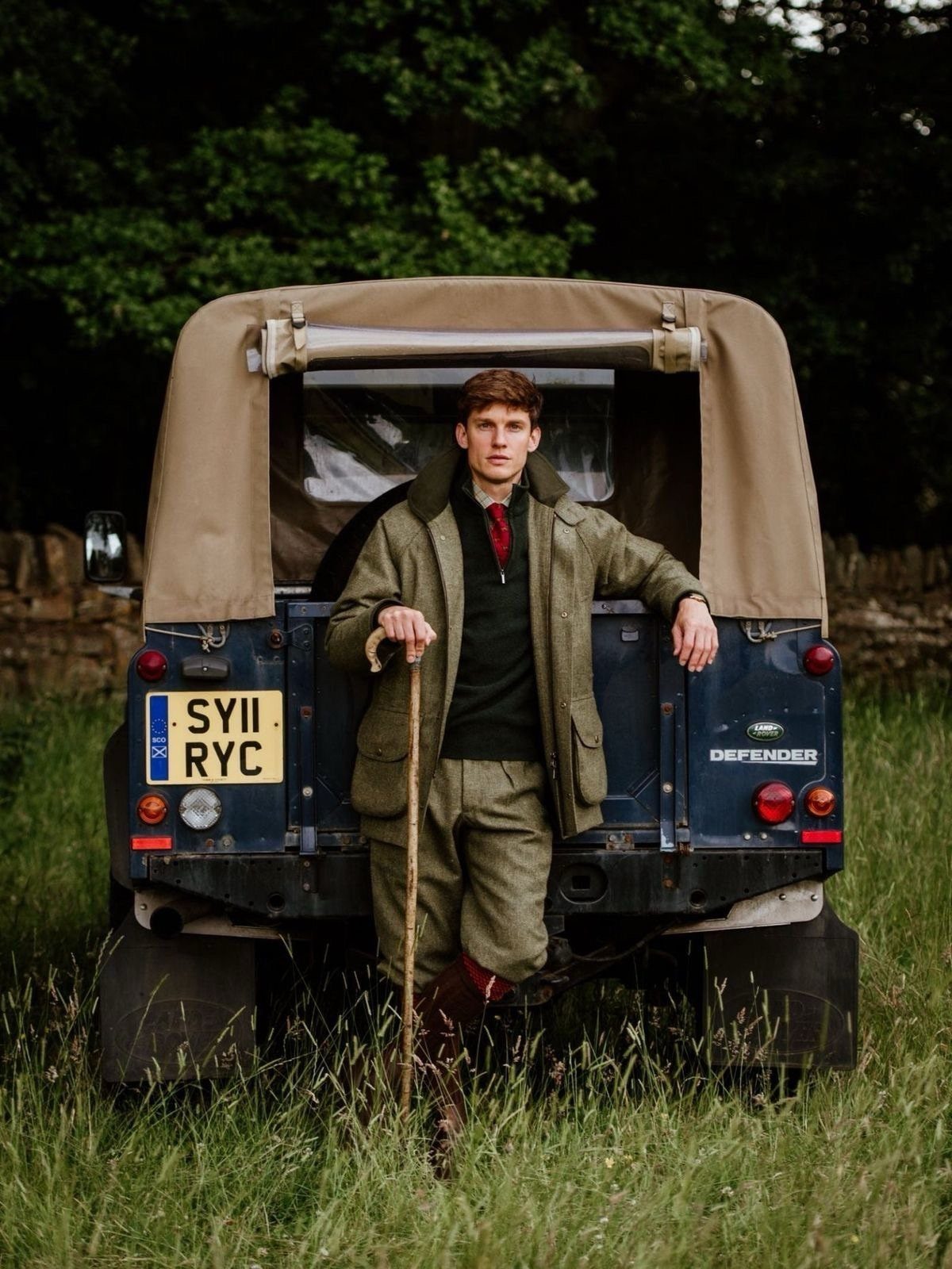 Man in country attire with brown boots standing before a blue off-road vehicle.