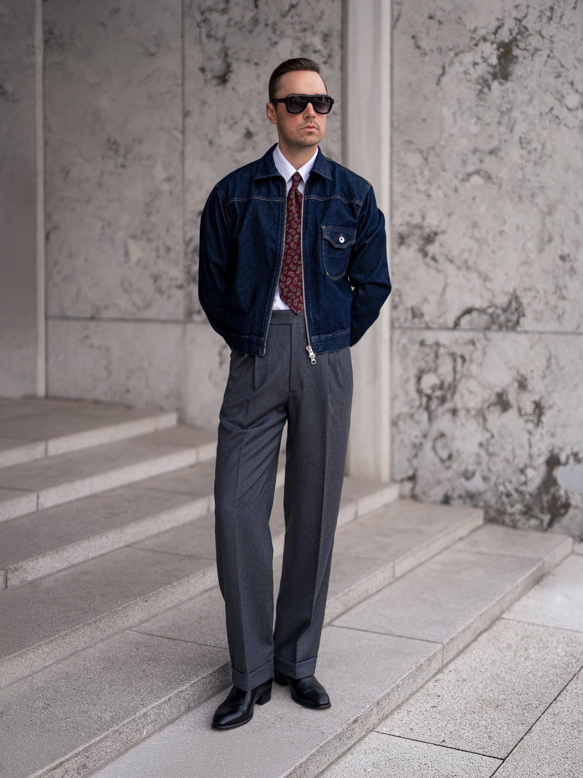 Man wearing denim jacket, tie, grey pleated trousers, and black leather boots.