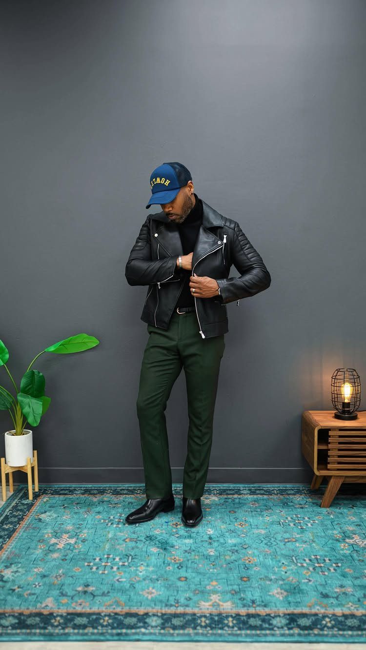 Man in leather jacket, turtleneck, and green pants wearing black boots on a rug.