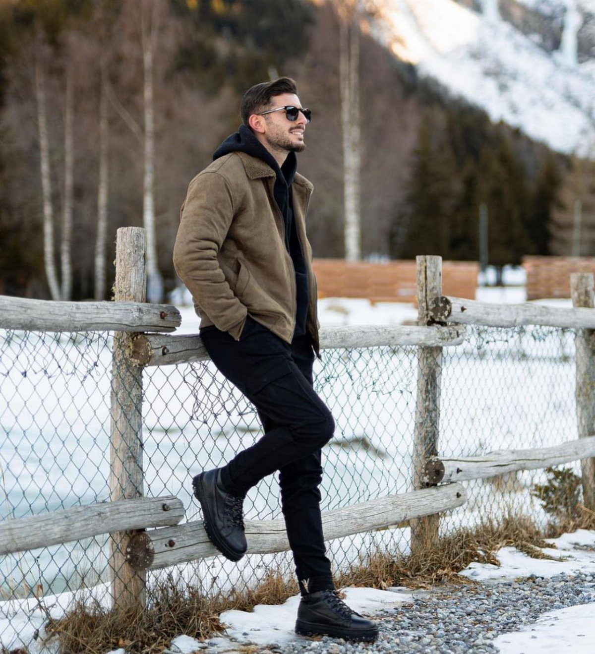 Man in brown jacket and black boots leaning on a fence in snow