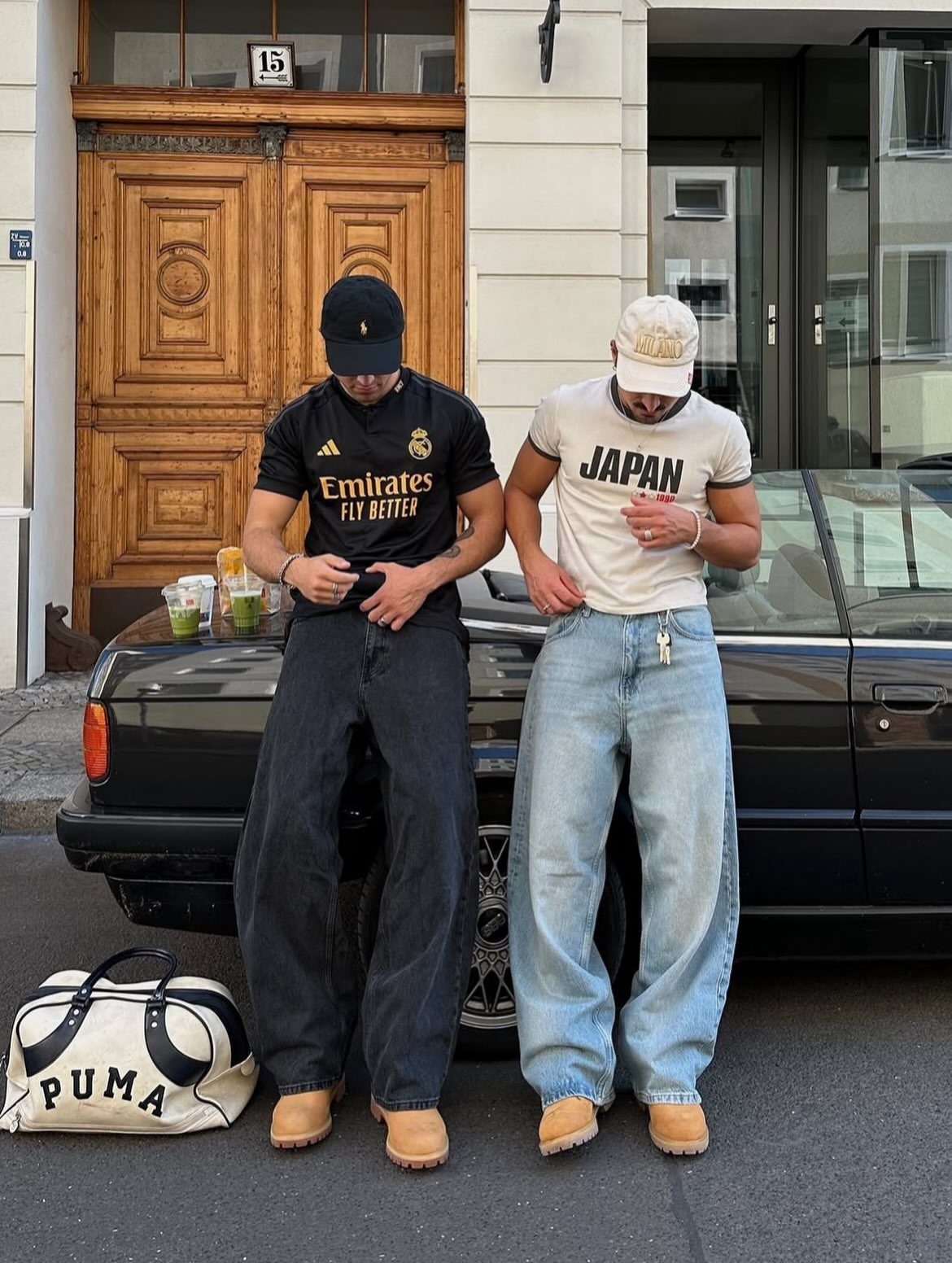 Two men in wide-leg jeans and tan boots leaning against a black car.
