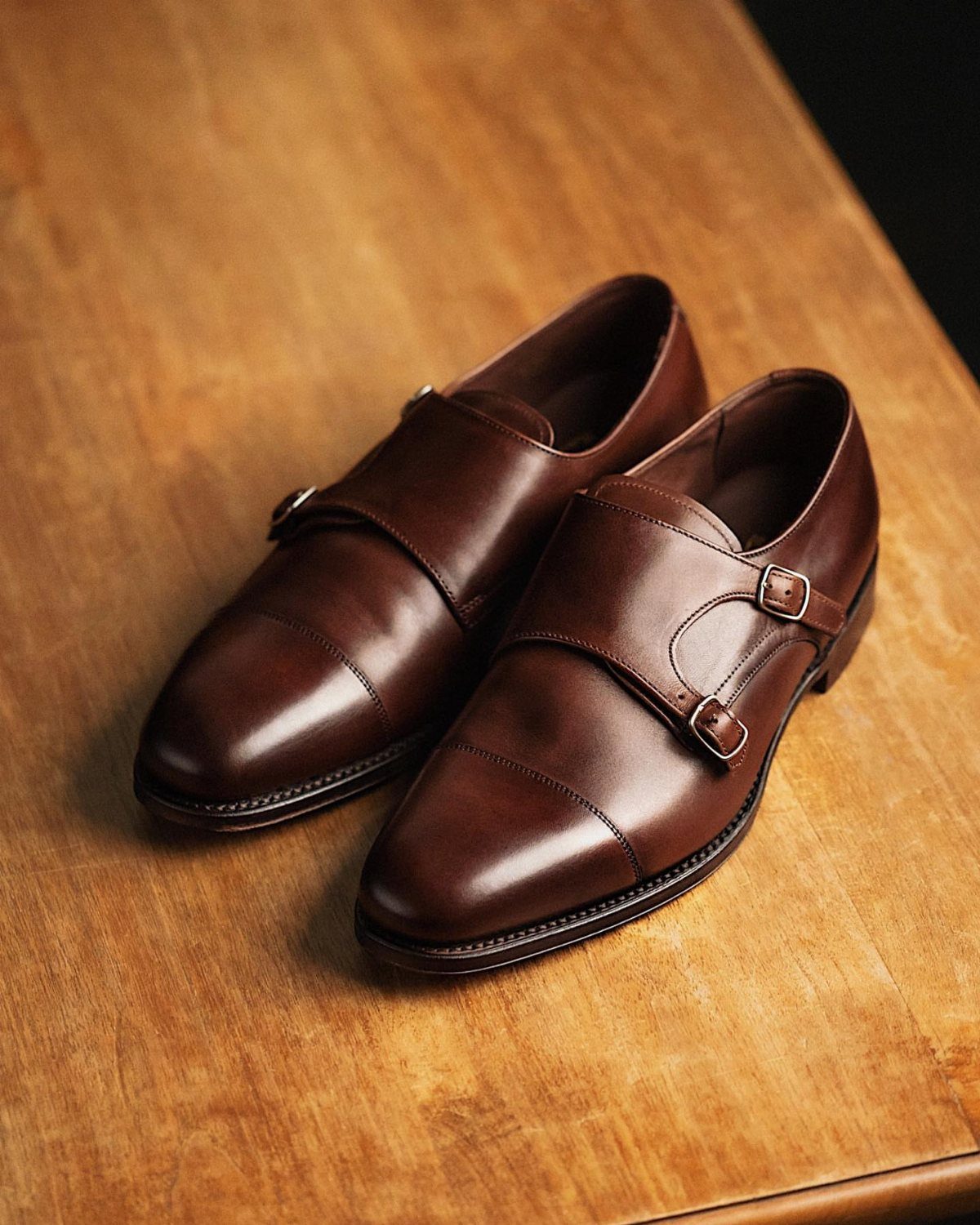 Brown leather double monk strap shoes on a light wooden table surface.