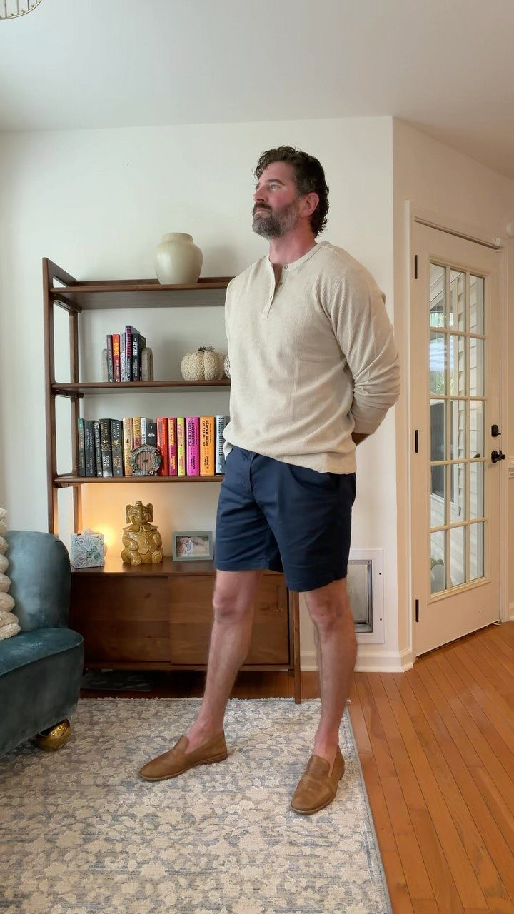 Man wearing beige henley, navy shorts, and brown suede loafers standing indoors.