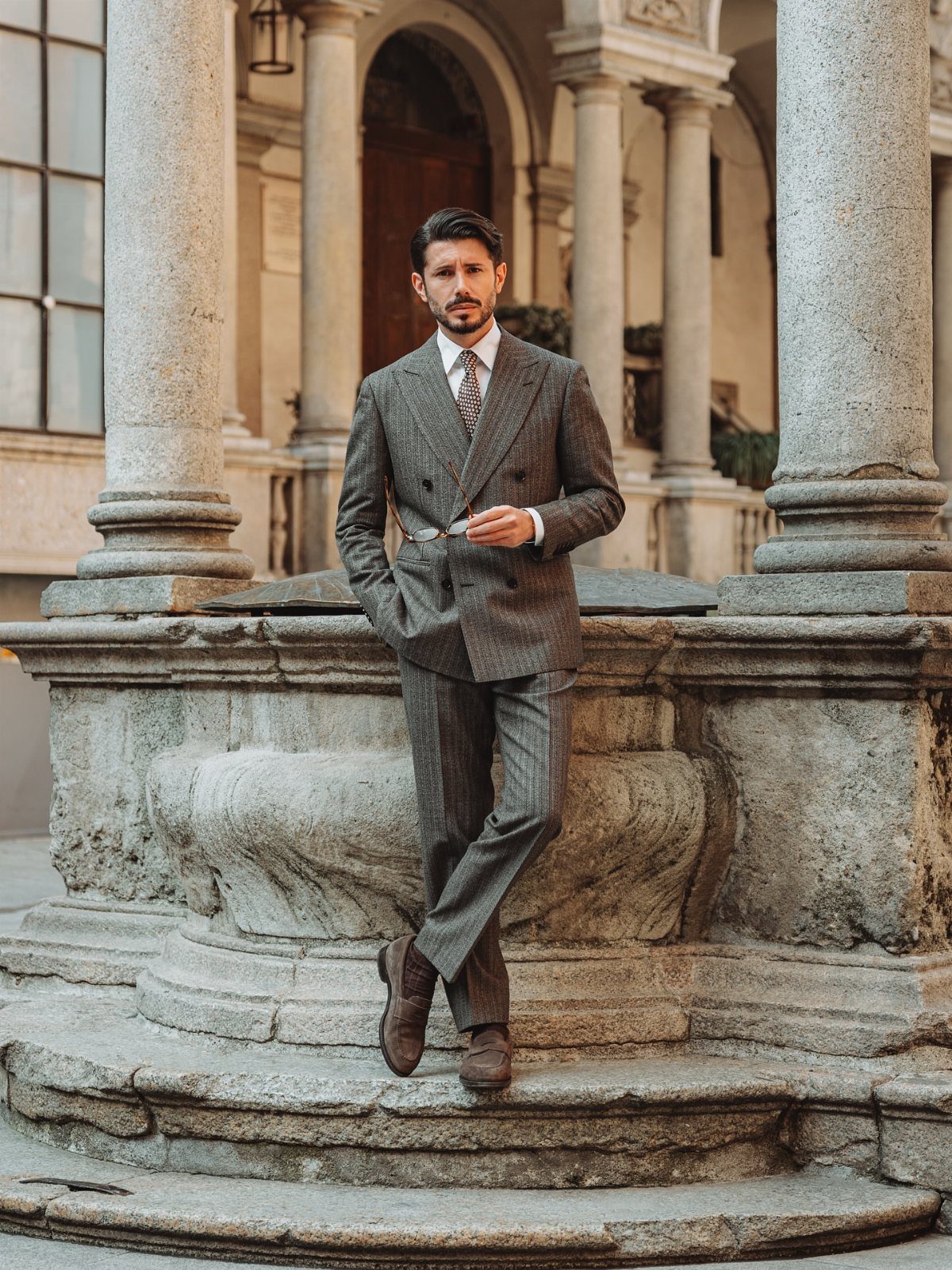 Man in a grey pinstripe suit and dark brown suede loafers leaning on stone.