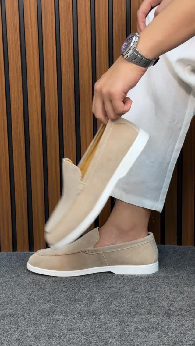 Light beige suede loafers paired with white chinos against a wooden wall background.