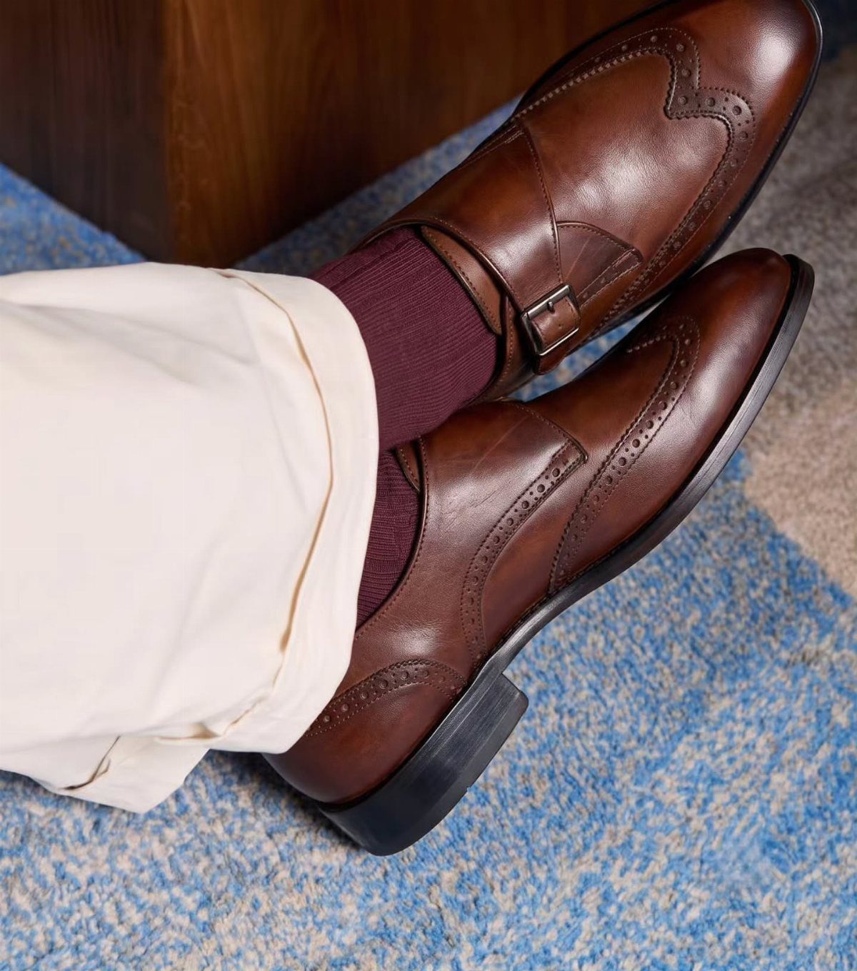 Brown leather monk strap shoes with beige trousers and burgundy socks on blue carpet.