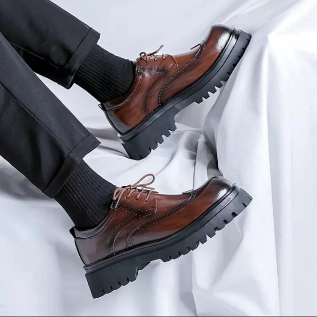 Brown leather dress shoes with chunky black soles and black trousers on white fabric.