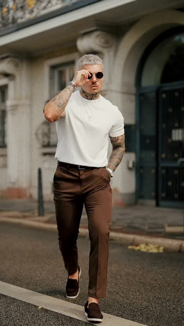 Man wearing white t-shirt, brown trousers, and dark brown loafers walking outdoors.
