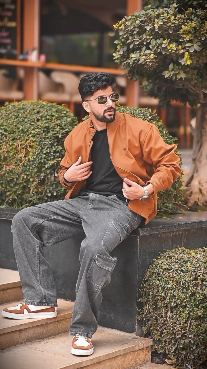 Man wearing brown sneakers, grey cargo pants, and a brown jacket sitting outdoors.