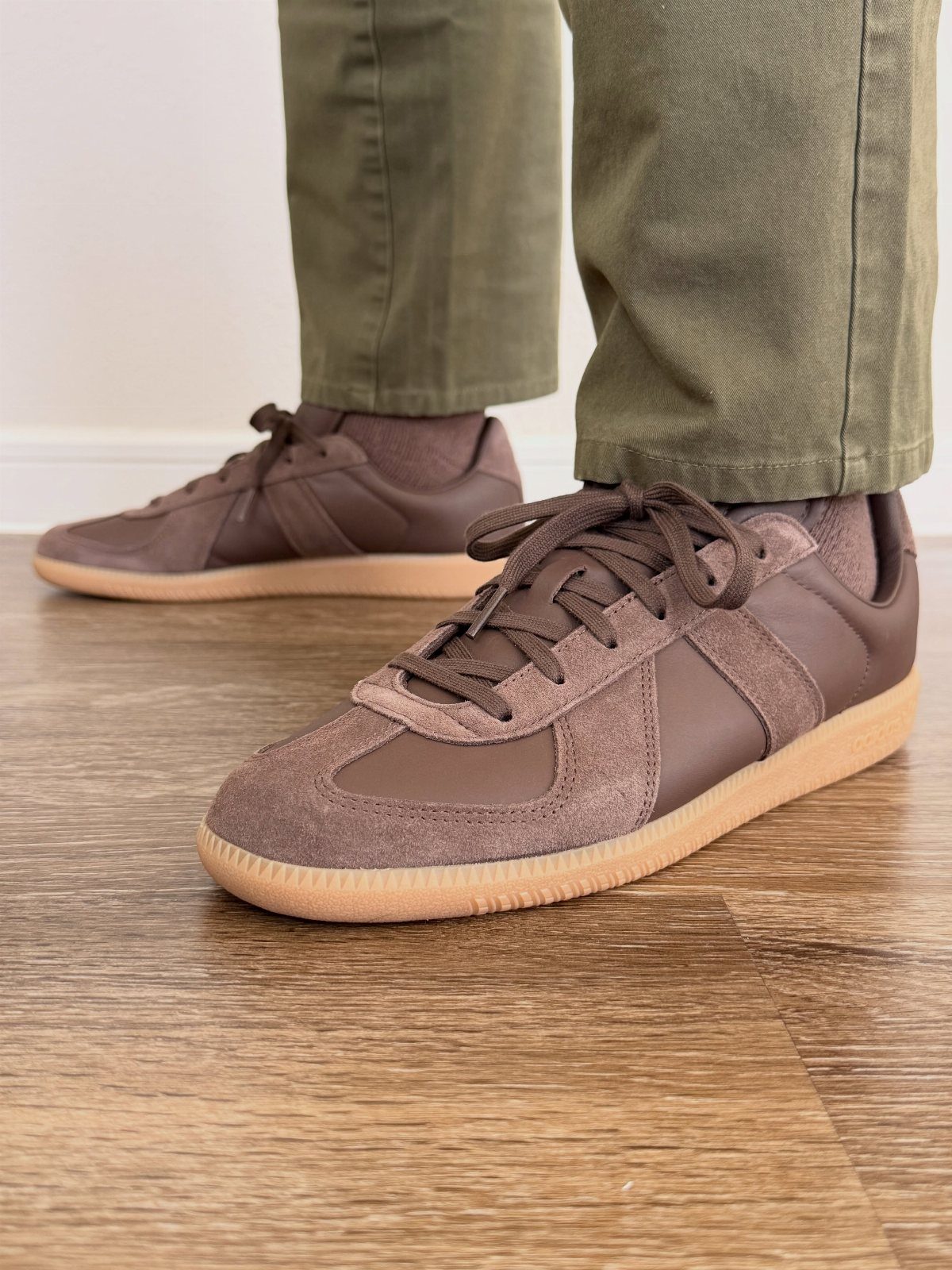 Brown suede sneakers with tan soles paired with olive green trousers on wooden floor