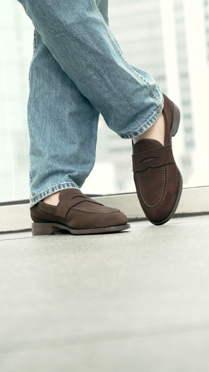 Brown suede loafers paired with light blue denim jeans on a concrete floor.