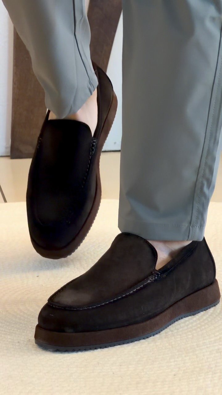 Dark brown suede loafers worn with light grey trousers on a white floor.