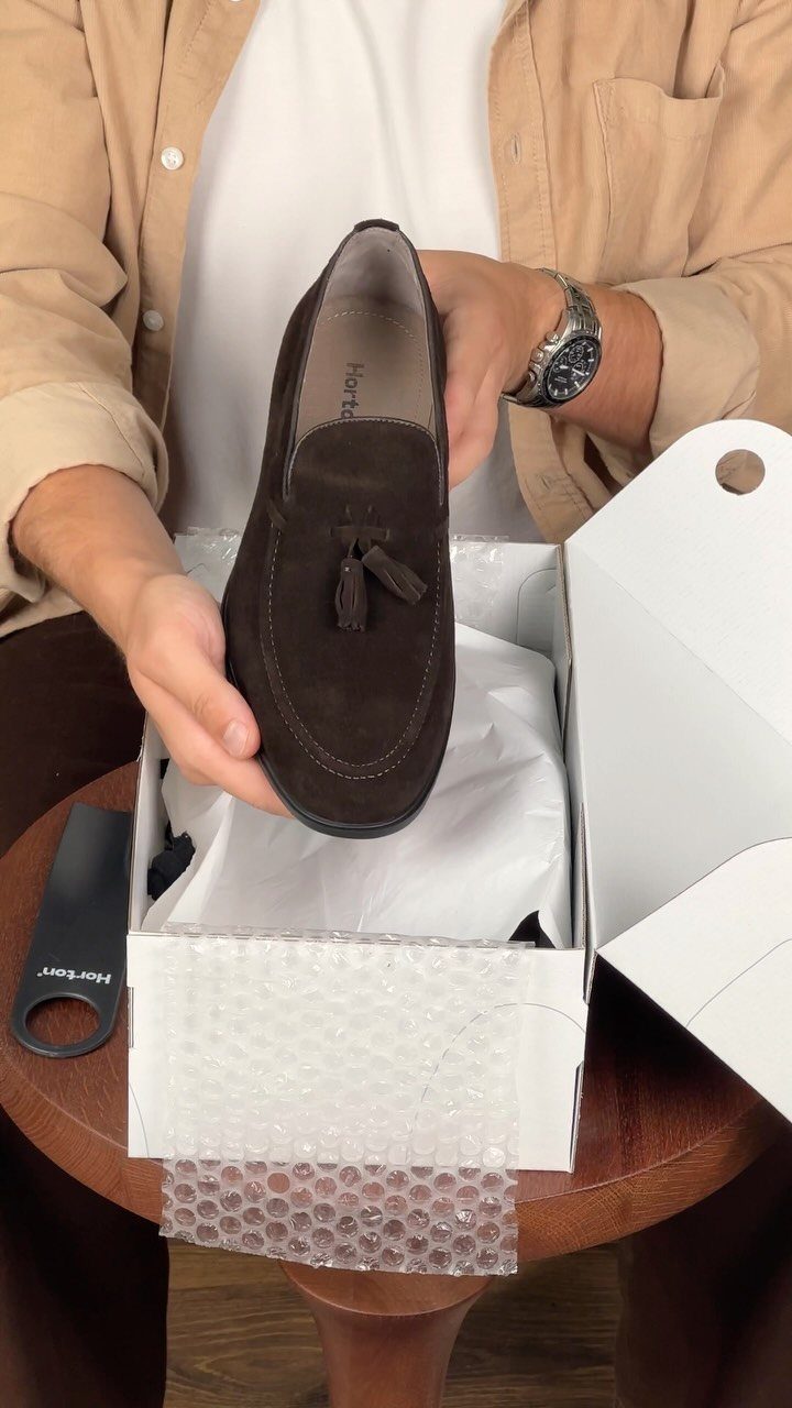 Brown suede tassel loafer held over a shoebox with a black shoe tree.