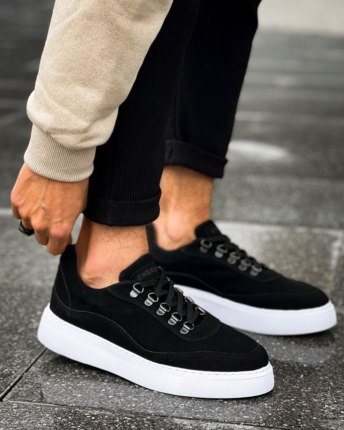 Black suede sneakers with white soles worn with black trousers and beige sweater.