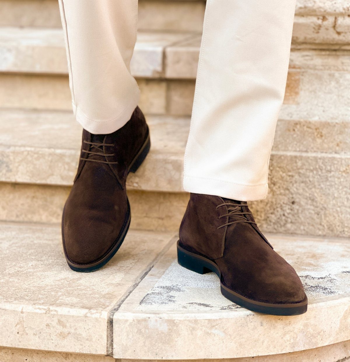 Dark brown suede chukka boots paired with off-white trousers on stone steps.