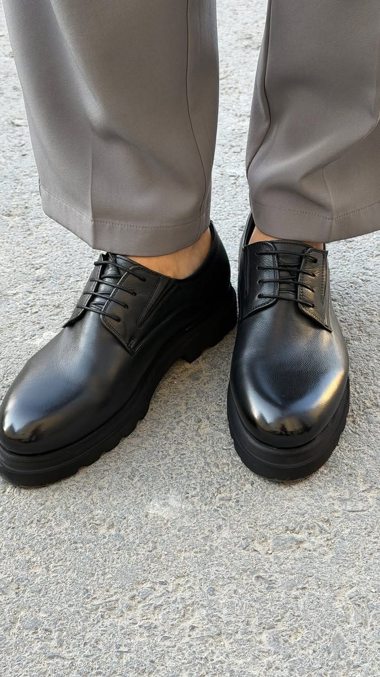 Black leather chunky derby shoes paired with grey trousers on concrete ground.